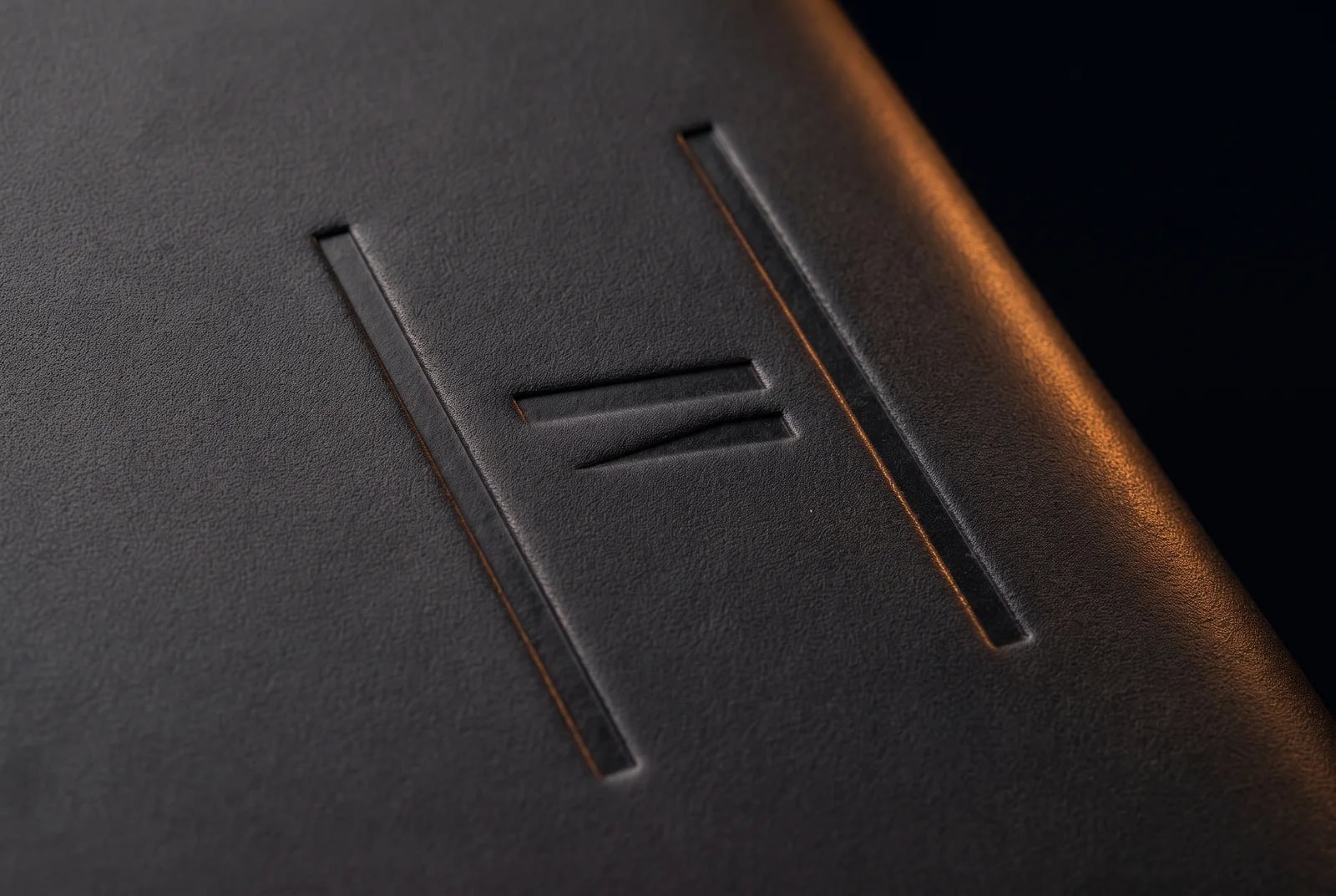 The HAUS:element monogram blind-debossed into dark leather, caught along a single seam of warm raking light.