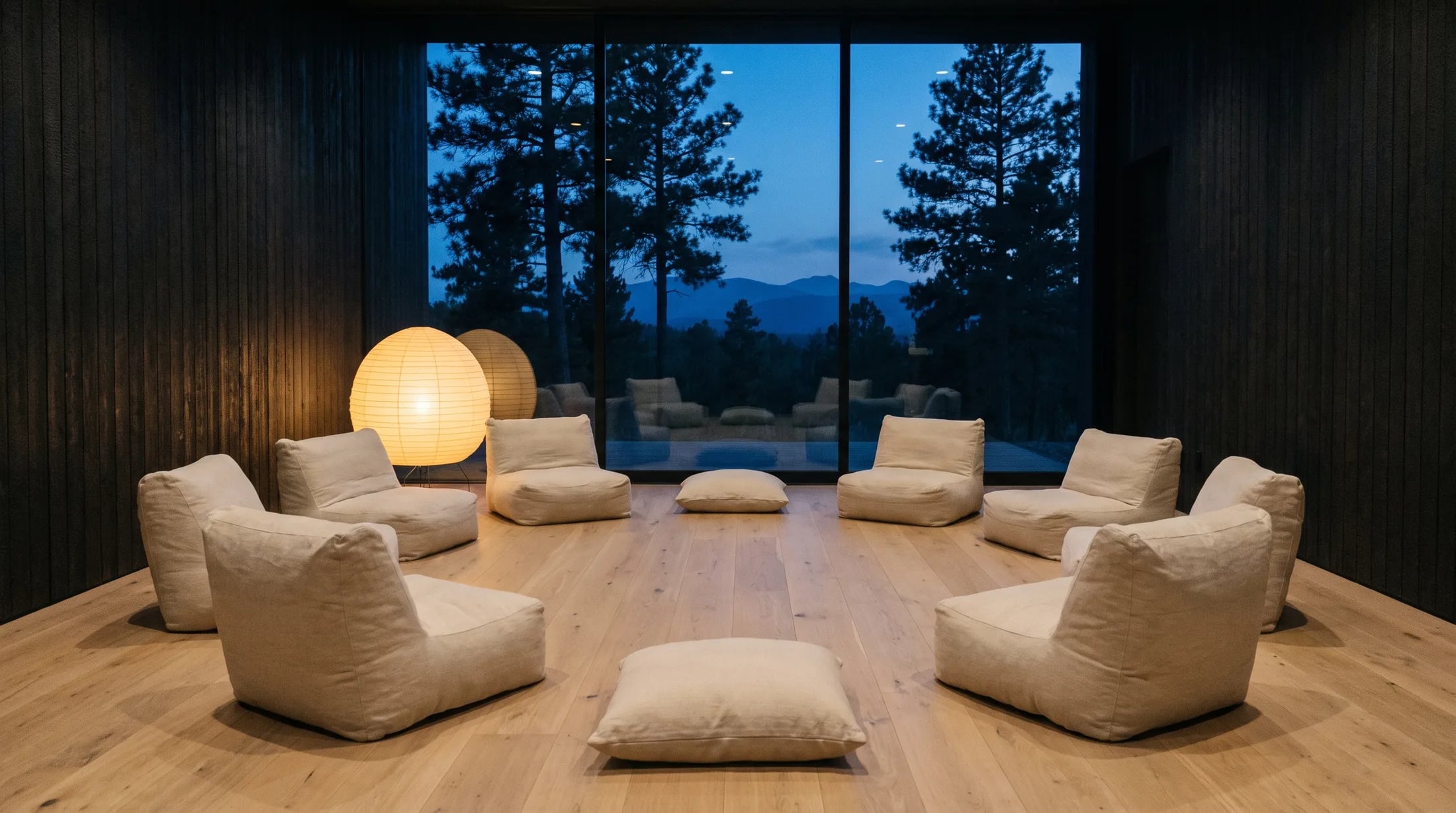 A low Japandi gathering hall at blue hour — dark timber, a loose circle of linen chairs on pale oak, a monumental window to black pines, one warm lamp glowing.