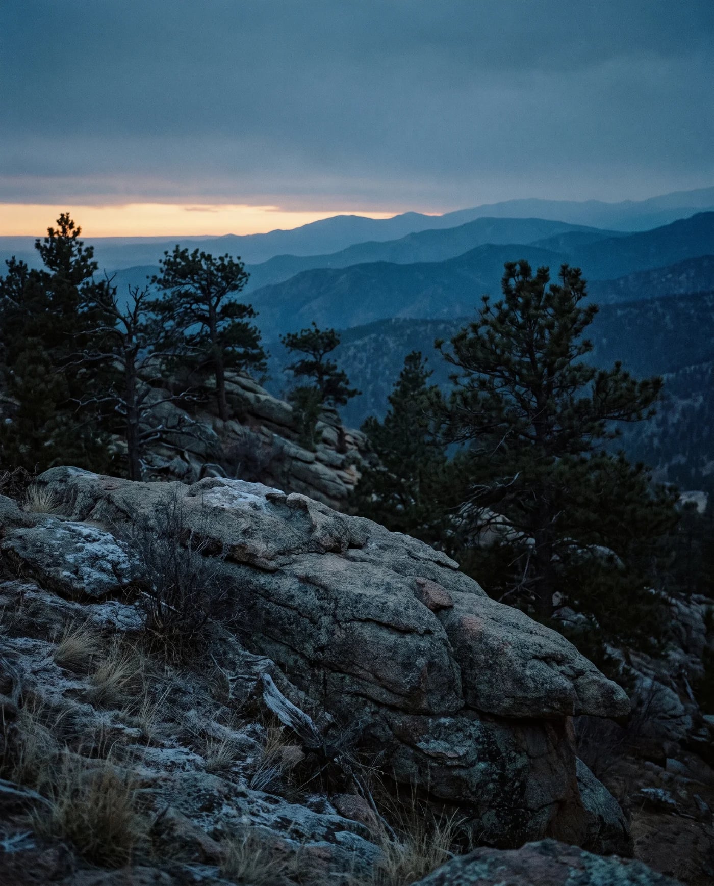 Raw Colorado foothill land at cold dusk — a weathered granite outcrop, scattered pines, ridgelines fading into haze, a sliver of pale amber last light.