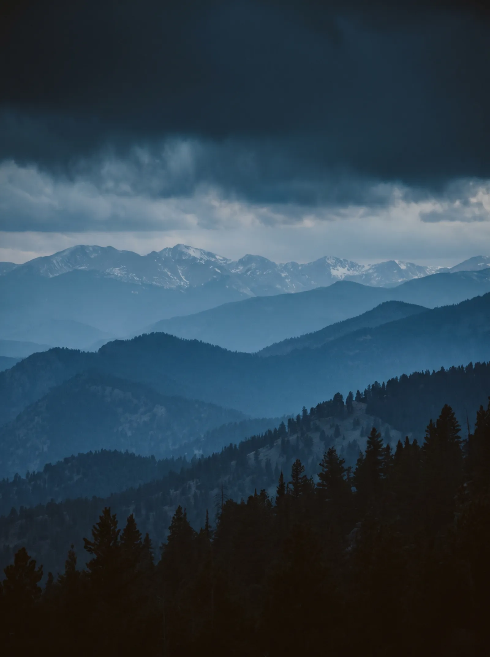 Layered mountain ridgelines receding into blue atmospheric haze at cold evening, distant snow-dusted peaks under a low storm sky.